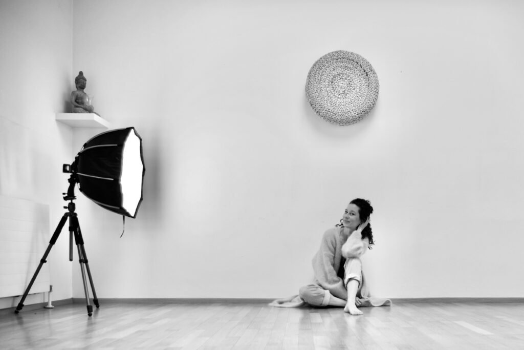 Eine Frau mit lockigem Haar sitzt gemütlich auf dem Boden eines hellen Raums, den linken Arm auf das aufgestelte linke Bein abgestützt, links steht ein Fotolicht auf einem Stativ, an der Wand hängen eine Buddha-Figur auf einem Regal und ein rundes Wandobjekt